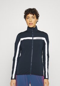 Giacca zip-up blu marina con strisce bianche lungo le maniche e logo sul petto. Collo alto, design aderente e maniche lunghe.