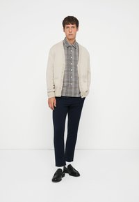Béžový pletený cardigan cez šedú a bielu károvanú košeľu s gombíkmi, v kombinácii s námorníckymi nohavicami a čiernymi mokasínami. Minimalistický dizajn, neformálny vzhľad.