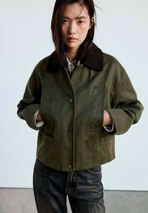 Mujer con el pelo liso y oscuro, vistiendo una chaqueta verde con cuello negro y vaqueros oscuros, de pie con las manos en los bolsillos de la chaqueta contra un fondo liso.