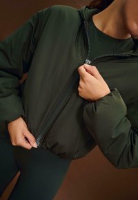 Zeleni puffer jakna s zatvaračem, sa visokim okovratnikom i prilagođenim manžetama. Materijal izgleda mekano i blago sjajno.