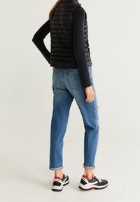 Gilet en duvet noir par-dessus un haut à manches longues, associé à un jean bleu clair et des baskets noires avec des accents rouges et blancs.