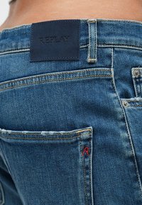 Σκούρο μπλε τζιν με υφή στην επιφάνεια, κίτρινο ράψιμο και δερμάτινο ταμπελάκι με την ένδειξη «REPLAY» στη μέση.