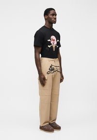 Μαύρο μπλουζάκι με πολύχρωμο γραφικό παγωτού, μπεζ παντελόνια cargo με σχέδιο γάτας, καφέ παπούτσια slip-on με σχέδιο. Λείο ύφασμα και casual εφαρμογή.