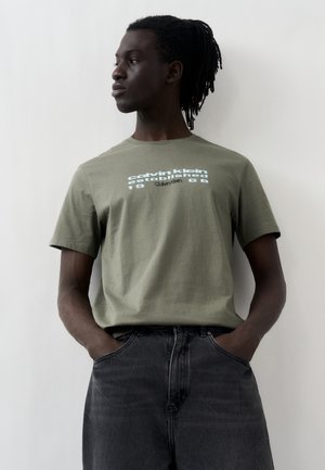 Jeune homme avec des dreadlocks portant un t-shirt Calvin Klein vert olive et un jean noir, debout les mains dans les poches, regardant vers la gauche.
