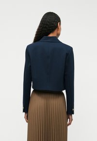 Kobieta z długimi kręconymi włosami, ubrana w skróconą granatową marynarkę i brązową spódnicę w plisy o wysokim stanie, stojąca plecami do kamery.