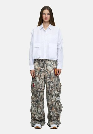 Jeune femme portant une chemise blanche oversize et un pantalon cargo ample avec un motif camouflage marron et gris et des patchs, debout devant un fond uni.