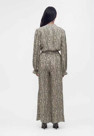 Femme aux longs cheveux foncés portant une combinaison longue manches, taille haute et jambes larges avec un imprimé animal beige et noir, vue de dos.