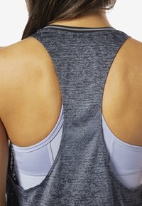 Šedý sportovní tílko s otevřeným zadním designem a texturovanou tkaninou. Ukazuje světlou fialovou sportovní podprsenku pod ním pro kontrast.