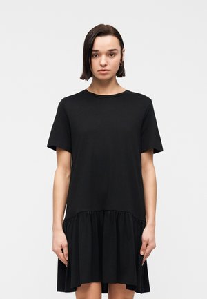 Woman with straight dark hair wearing a plain black short-sleeve dress with a gathered hem standing against a white background.