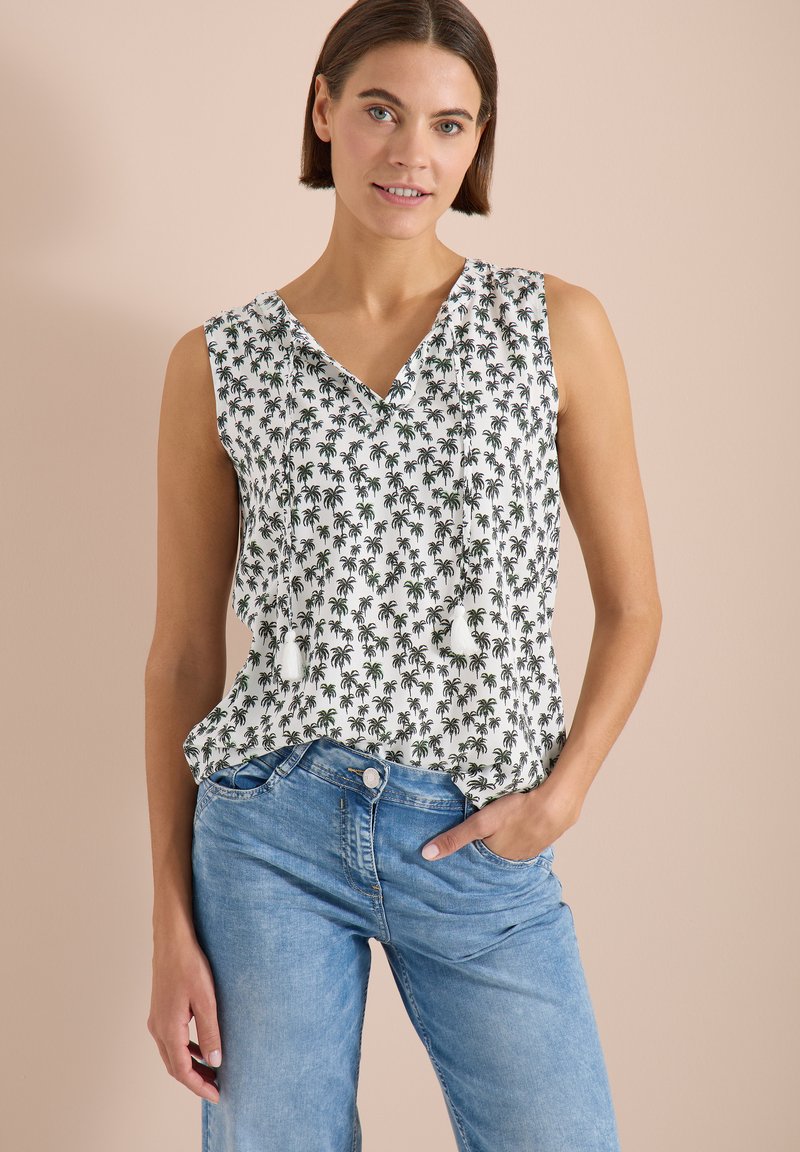Femme portant un haut blanc sans manches à col en V avec imprimé de palmiers noirs et liens à pompons, associée à un jean bleu clair, debout contre un mur beige.