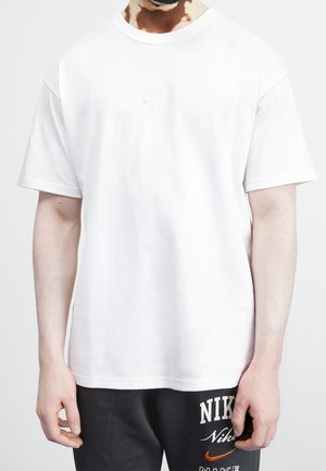 Personne portant un t-shirt blanc uni Nike et un short noir Nike avec logos visibles, debout devant un fond clair uni.
