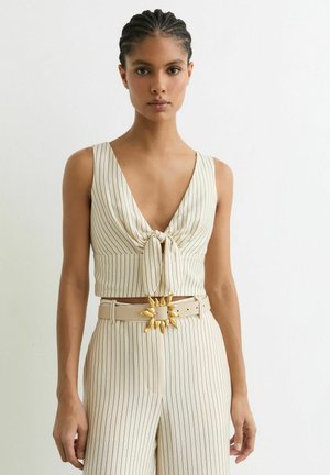 Kobieta z warkoczami, ubraną w kremowy, bezrękawowy krótki top z pionowymi paskami, związany z przodu, oraz dopasowane, wysokie spodnie z paskiem z złotą klamrą.