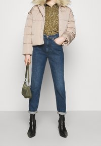 En beige puffjakke med pelskrave, mønstret grøn bluse, højttaljede mørkeblå jeans og sorte ankelstøvler. Bærer en grøn taske.