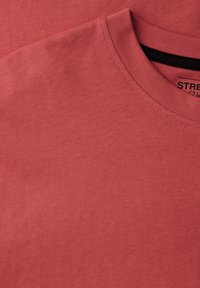 Gros plan sur un t-shirt en coton couleur rose poudré, à col rond, plié, avec des coutures visibles autour du col et une étiquette noire partielle à l'intérieur.