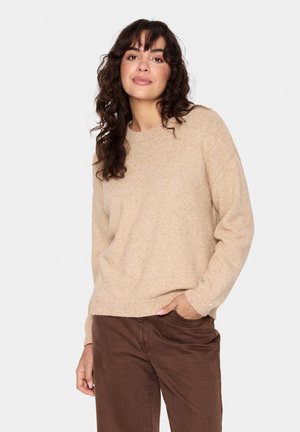 Bežinis megztinis su tekstūruotu mezginiu, apvaliu kaklu ir žinoti rankogaliais, derinamas su rudomis kelnėmis. Šviesios dėmės suteikia vizualinį susidomėjimą.