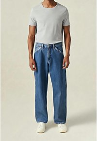 Homme portant un t-shirt gris clair à manches courtes, un jean bleu ample et des chaussures blanches, debout devant un fond uni.