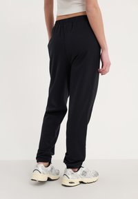 Sorte joggebukser med elastisk midje, sidelommer og tapered ben, kombinert med sølvfargede joggesko. Materialet virker mykt og elastisk.