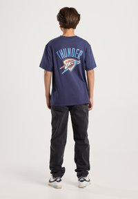 Niño de pie de espaldas, vistiendo una camiseta azul marino de Thunder OKC, jeans negros y zapatillas Nike blancas sobre un fondo liso.