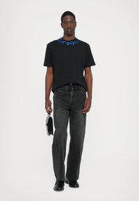 Svart t-shirt med blå text vid halsringningen, kombinerad med svarta denimjeans med kontrasterande sömmar i sidan och svarta skor.