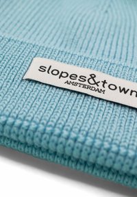Jasnoniebieska dziana czapka z prążkowaną teksturą. Na przedniej części znajduje się biały metek z czarnym napisem "slopes & town AMSTERDAM".