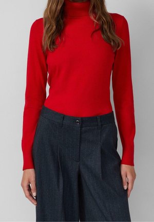 Vrouw draagt een aansluitende rode coltrui die is ingestopt in hoge taille, donkergrijze wijde broek met krijtstreep.