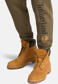 Braune Timberland-Stiefel und olivgrüne Jogginghosen mit "Timberland"-Logo, Fokus auf Fußbekleidungsdetails und Stofftextur der Kleidung.