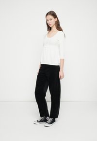 Λευκό πλεκτό τοπ με μανίκια τριών τετάρτων και λεπτομέρεια με κουμπιά, συνδυασμένο με χαλαρά μαύρα παντελόνια κοτλέ και μαύρα αθλητικά παπούτσια.