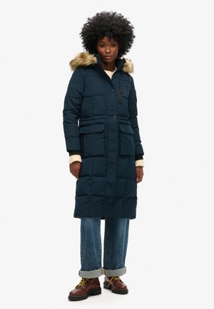 Marineblauwe gewatteerde parka met een nepbont capuchon, ritssluiting en knoopsluiting, twee voorkantzakken en een middellange design, gecombineerd met opgerolde jeans en laarzen.
