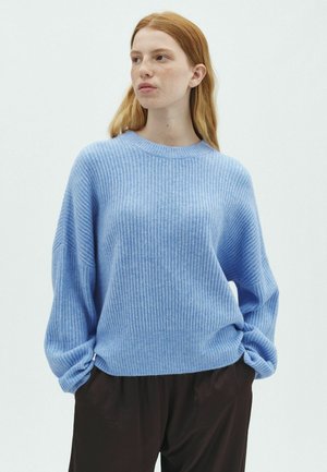 Jonge vrouw met rood haar, die een lichtblauwe geribbelde trui en donkere losse broek draagt, staand met handen in de zakken tegen een witte achtergrond.