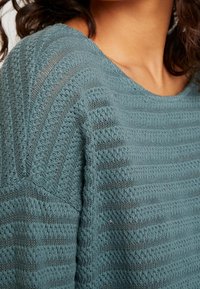 Maglione in maglia teal con strisce testurizzate; presenta un collo rotondo e modelli a punto aperto per una maggiore traspirabilità. Finitura morbida e vestibilità rilassata.