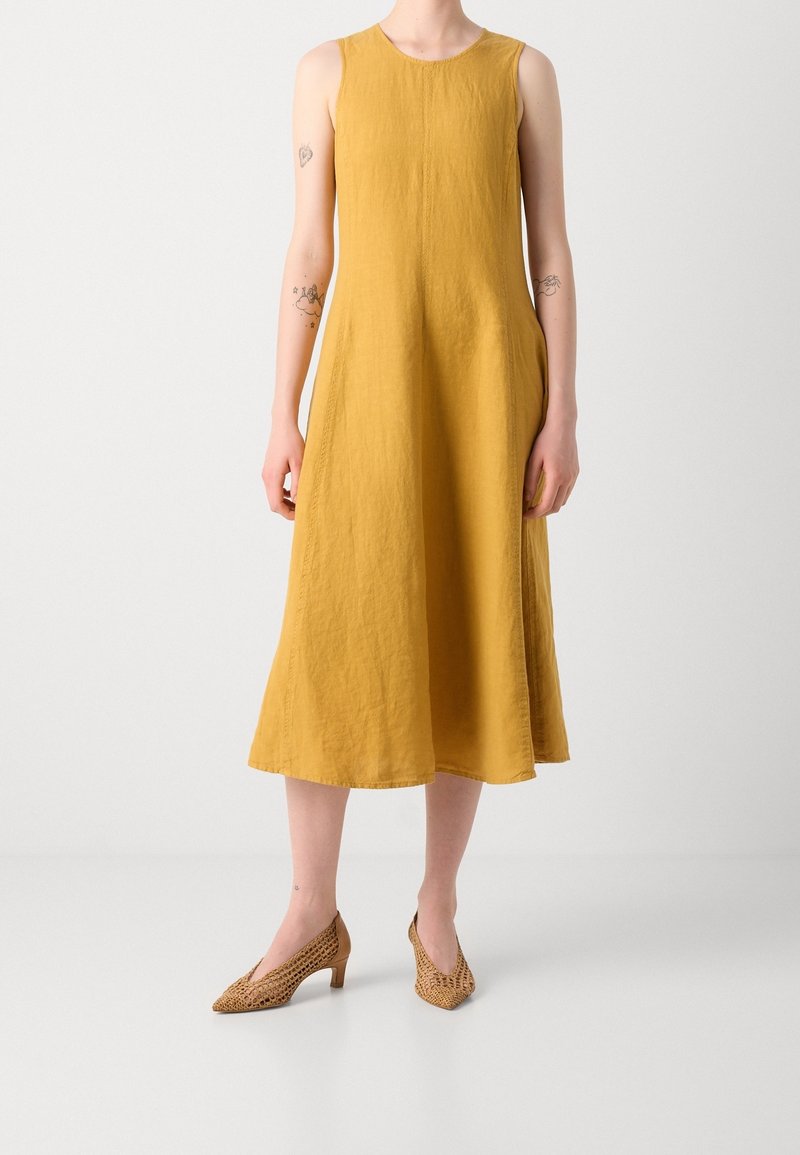 Robe midi sans manches jaune moutarde portée avec des chaussures tissées marron clair à talon bas, montrée des épaules aux pieds sur un fond blanc uni.