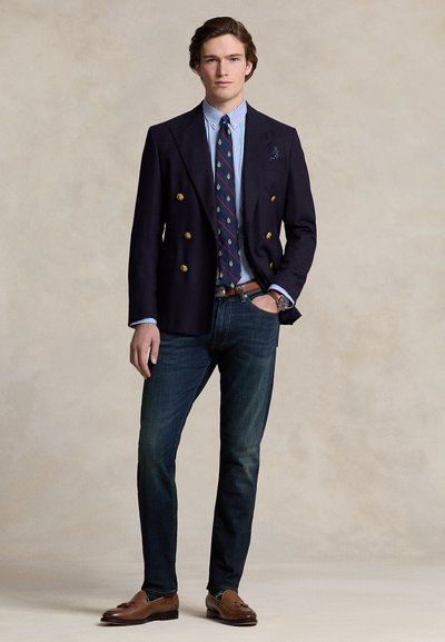 Tmavomodrý obojstranný blejzer so zlatými gombíkmi, pásikovaná košeľa, vzorovaná kravata, tmavé džínsy a hnedé mokasíny. Hladká textúra, na mieru šit ý strih.