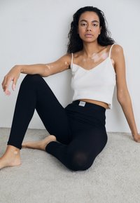 White ribbed tank top with lace trim, paired with black leggings featuring a logo waistband. Model seated on a soft, textured surface.
