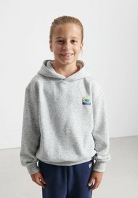 Felpa grigia in pile con tasca frontale, cappuccio regolabile con cordino e una patch colorata sul petto, abbinata a pantaloni della tuta blu navy.