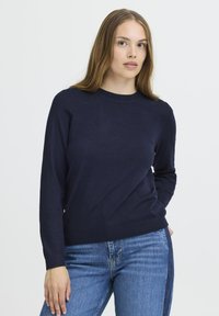 Pull en maille bleu marine avec un col rond et des manches longues, présentant une texture lisse et des poignets et un ourlet côtelés. Associé à un jean bleu.