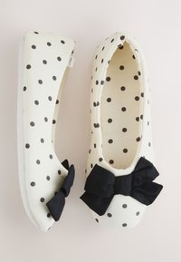 Chaussons blancs slip-on avec des pois noirs et de grands nœuds en tissu noir à l'avant, présentés sur un fond neutre.