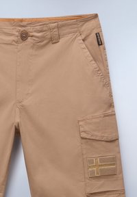 Beige cargo bukser med knaplukning, sidelommer og et dekorativt patch med et flagdesign på låret. Strukturstof.