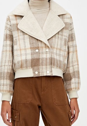 Femme portant une veste cropped à carreaux beige et marron sur un col roulé crème, associée à un pantalon cargo marron taille haute avec de grandes poches.