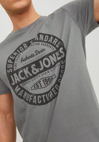 Uomo che indossa una maglietta grigia con una grande grafica circolare nera con la scritta "JACK & JONES Superior Standard Manufactured Goods EST 1990."