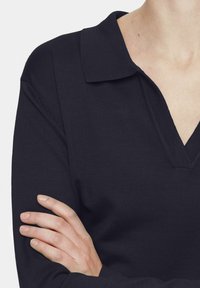 Navy ribbed knit top with a v-neck and a collar, featuring textured fabric. Arm crossed, showing a close-up of the garment.
