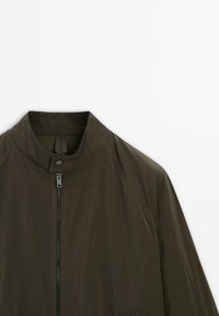 Donkergroene olijfgroene jas met hoge kraag, ritssluiting en een gladde textuur. Voorzien van raglanmouwen en een minimalistisch ontwerp.