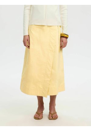 Osoba nosí bledožltú midi zavinovaciu sukňu, biele tričko s dlhým rukávom, hnedý náramok a béžové žabky na jednofarebnom pozadí.