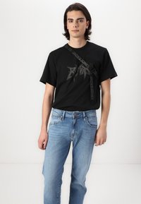 Black short-sleeve t-shirt features a prominent gray graphic and a logo strap. Paired with light blue denim jeans with a classic fit.
