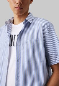 Camicia a maniche corte a righe blu e bianche, realizzata in tessuto leggero, con colletto button-down e una tasca sul petto.