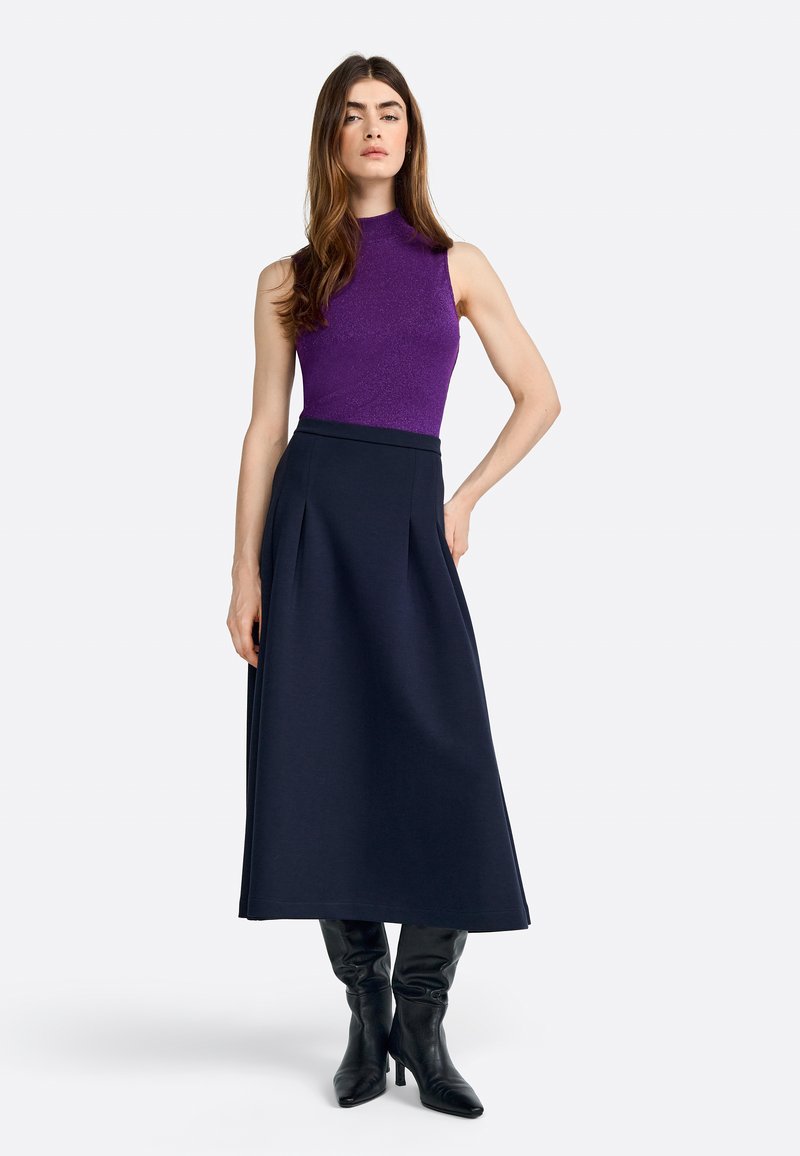 Top sans manches ajusté violet associé à une longue jupe plissée marine. Le mannequin porte des bottes en cuir noir hautes, se tenant devant un fond uni.