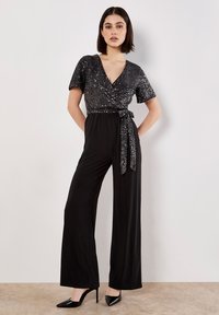 Vrouw in een zwarte jumpsuit met een glinsterende wrap-top en wijde broekspijpen, poseert met handen in de zakken tegen een witte muur.