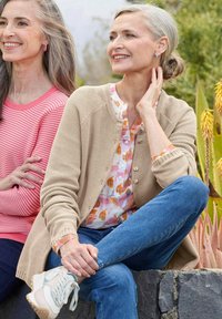 Gilet en maille beige sur un blouse à motifs floraux, associé à un jean bleu et des baskets blanches. Textures douces, couleurs chaudes et coupe décontractée.