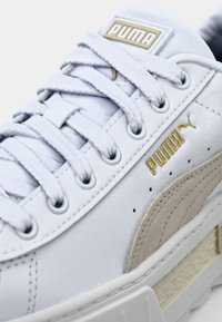 Weiße Sneaker mit einer strukturierten Lederoberfläche, versehen mit goldenen Akzenten, auffälligen Schnürsenkeln und einem markanten, tonalen Seitenpaneel-Design.