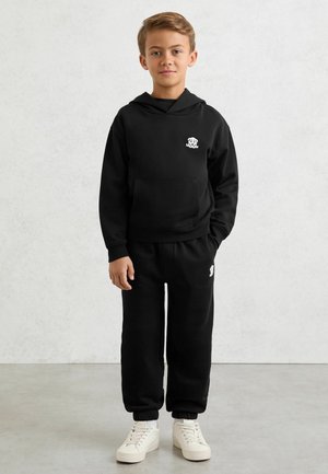 Mladý chlapec stojí, má na sobě černou mikinu s kapucí a slavnostní tepláky se stejnými bílými logy a bílé tenisky na jednoduchém pozadí.