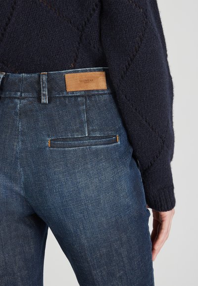 Vaqueros de mezclilla azul oscuro con una etiqueta de cuero en la parte trasera de la cintura, con un corte ajustado y un bolsillo trasero con detalle de costura horizontal.